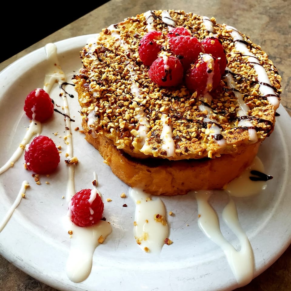 Thick French toast topped with chopped nuts, raspberries, white icing, and chocolate drizzle on a white plate.