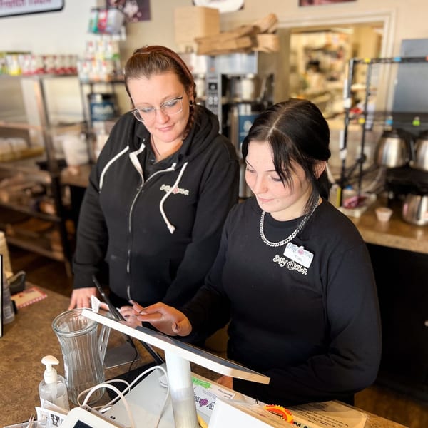 Two Sassy Onion employees, one training the other on a point-of-sale system at the counter.