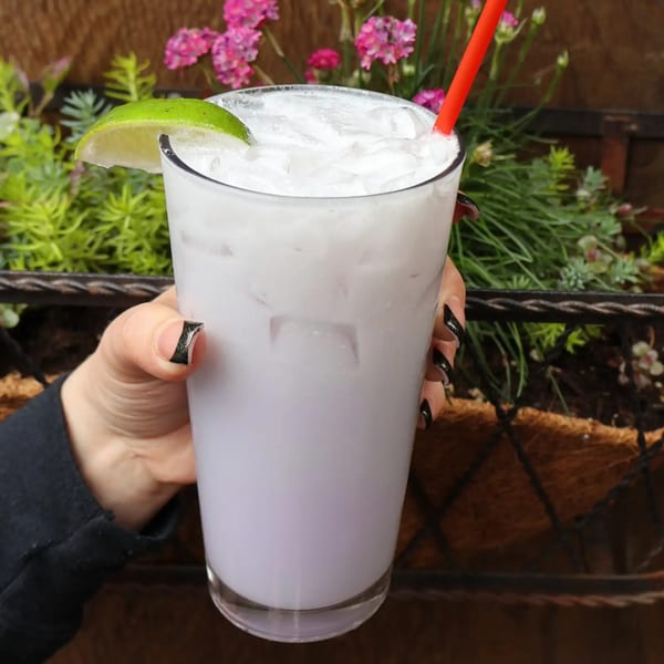 A hand holding a Sassy Onion cocktail with a lime wedge and red straw, against a floral background.