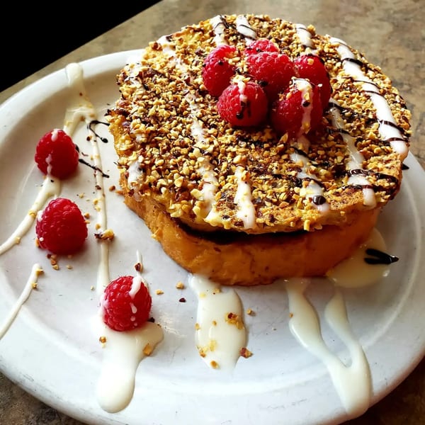 Thick French toast topped with raspberries, chopped nuts, white icing, and chocolate drizzle on a white plate.