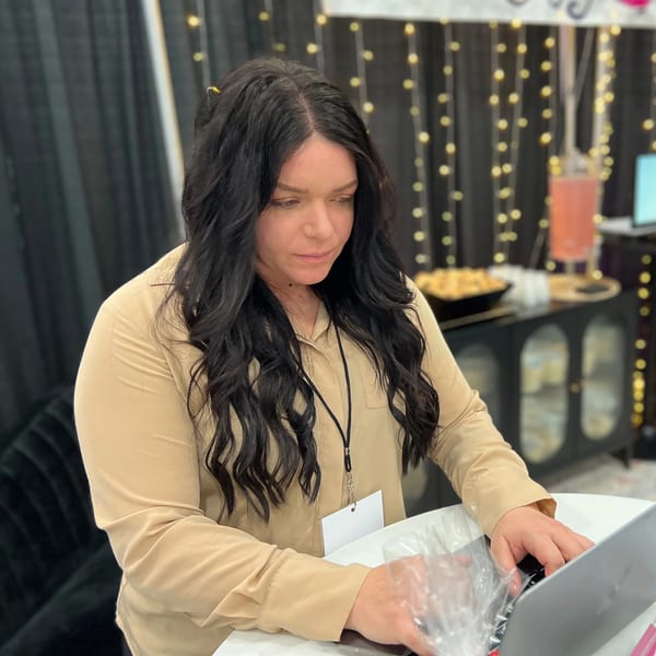 A woman with long dark hair in a tan shirt works on a laptop at an event.