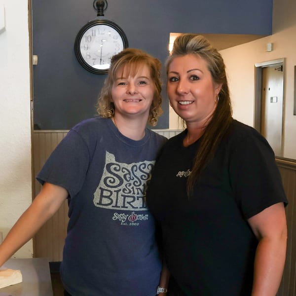 Two smiling women, one in a Sassy Onion t-shirt, stand in a restaurant with a clock.