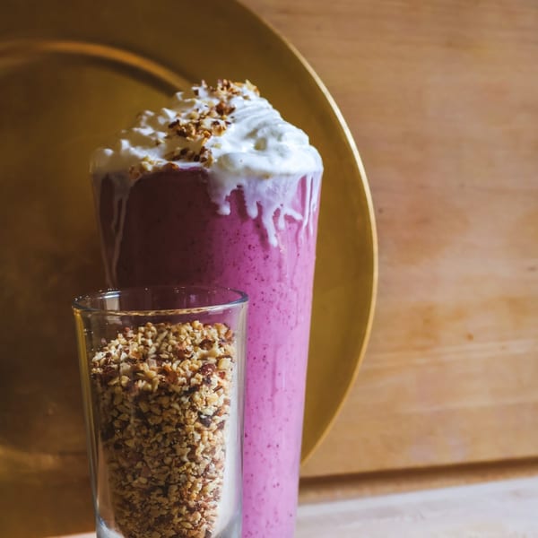A vibrant pink smoothie with whipped cream and chopped nuts, next to a glass of chopped nuts.